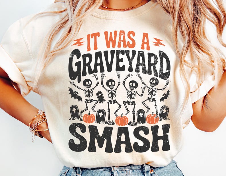 May include: Cream-colored t-shirt with a Halloween-themed graphic. The design features the text "IT WAS A GRAVEYARD SMASH" in orange and black, along with dancing skeletons, pumpkins, and tombstones. A fun and festive design.