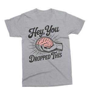 May include: A light gray t-shirt with the text "Hey, You Dropped This." The graphic features a pink brain held in a hand, with radiating lines. The design is in black and white.