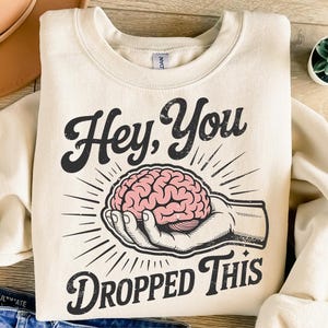 May include: Cream-colored sweatshirt with the text "Hey, You Dropped This" in a vintage font. The graphic features a pink brain held in a hand, with radiating lines. The sweatshirt is folded on a wooden surface.