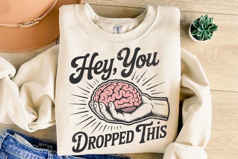 May include: Cream-colored sweatshirt with the text "Hey, You Dropped This" in a vintage font. The graphic features a pink brain held in a hand, with radiating lines. The sweatshirt is folded on a wooden surface.