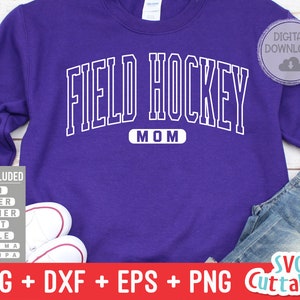 May include: Purple sweatshirt with the text "FIELD HOCKEY MOM" in white outlined letters. The sweatshirt is on a wooden surface with a pair of white sneakers and blue jeans.