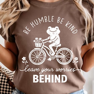 May include: Brown t-shirt featuring a white illustration of a frog on a bicycle, with a basket of flowers. The text says "BE HUMBLE BE KIND" and "leave your worries BEHIND".