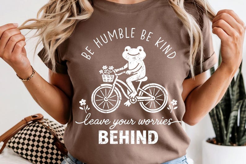 May include: Brown t-shirt featuring a white illustration of a frog on a bicycle, with a basket of flowers. The text says "BE HUMBLE BE KIND" and "leave your worries BEHIND".