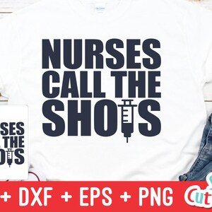 Pu&ograve; includere: Una maglietta bianca con il testo "NURSES CALL THE SHOTS" in lettere blu scuro. Una siringa &egrave; incorporata nella parola "SHOTS". L'immagine mostra anche una tazza bianca con lo stesso testo e un paio di jeans blu.