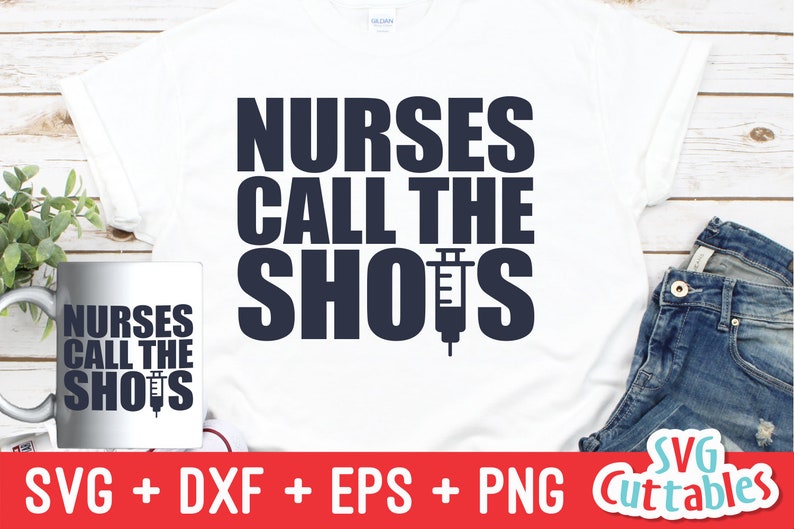 Pu&ograve; includere: Una maglietta bianca con il testo "NURSES CALL THE SHOTS" in lettere blu scuro. Una siringa &egrave; incorporata nella parola "SHOTS". L'immagine mostra anche una tazza bianca con lo stesso testo e un paio di jeans blu.