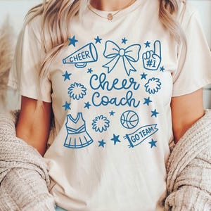May include: A beige t-shirt with a blue graphic design featuring the words "Cheer Coach" surrounded by cheerleading-related icons. The icons include a megaphone, bow, pom-poms, a basketball, and a cheerleader uniform.