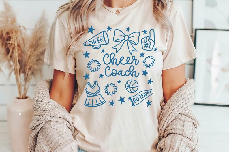 May include: A beige t-shirt with a blue graphic design featuring the words "Cheer Coach" surrounded by cheerleading-related icons. The icons include a megaphone, bow, pom-poms, a basketball, and a cheerleader uniform.