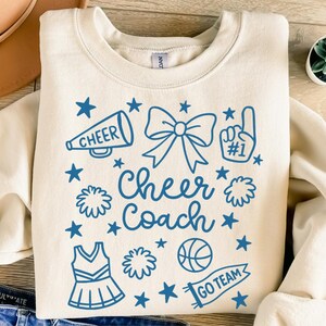 May include: Cream-colored sweatshirt with a blue graphic that says "Cheer Coach." The design includes a megaphone, bow, pom-poms, stars, a cheerleader uniform, a basketball, and a "Go Team" banner.