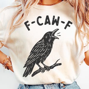 May include: Cream-colored t-shirt featuring a black graphic of a crow perched on a branch, with the text "F-CAW-F" above the bird. The crow is depicted with its beak open. The shirt has short sleeves.