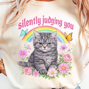 Pu&ograve; includere: T-shirt color crema con illustrazione di gatto tigrato grigio e la scritta "silently judging you". Il gatto &egrave; circondato da fiori, un arcobaleno e farfalle. Il design &egrave; stravagante e colorato.