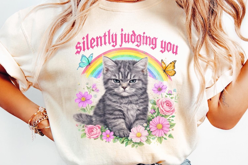 Pu&ograve; includere: T-shirt color crema con illustrazione di gatto tigrato grigio e la scritta "silently judging you". Il gatto &egrave; circondato da fiori, un arcobaleno e farfalle. Il design &egrave; stravagante e colorato.
