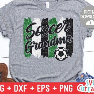 May include: Gray t-shirt with the words "Soccer Grandma" in black script over green, white, and black brushstrokes. A soccer ball graphic is below the text. The words "Digital Download" are in a white circle.