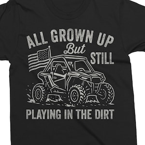 May include: Black t-shirt with a white graphic of an off-road vehicle, an American flag, and the text "ALL GROWN UP But STILL PLAYING IN THE DIRT". The design has a vintage, distressed look.
