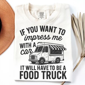 May include: Cream-colored t-shirt with the text "IF YOU WANT TO impress me WITH A car IT WILL HAVE TO BE A FOOD TRUCK" in black. The graphic includes a food truck illustration.