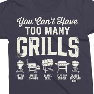 May include: Dark blue t-shirt with the text "You Can't Have Too Many Grills" in white. Below the text are illustrations of different grill types, including kettle, offset smoker, barrel, flat top griddle, and classic backyard grill.