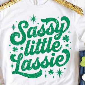 May include: White t-shirt with the text "Sassy little Lassie" in green, decorated with shamrocks and stars. The shirt is displayed on a gold sequined hanger, with shamrock and gold coin decorations.