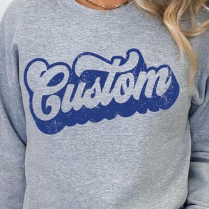 May include: A gray sweatshirt with the word "Custom" in a retro blue font. The word has a distressed, vintage look, with a white outline. The sweatshirt is a casual style, suitable for everyday wear.