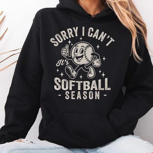 Op de afbeelding: Zwarte hoodie met een softbalafbeelding. De afbeelding toont een softbalpersonage met een duim omhoog en de tekst "SORRY I CAN'T It's SOFTBALL SEASON". De tekst en afbeelding zijn in wit en lichtgrijs.