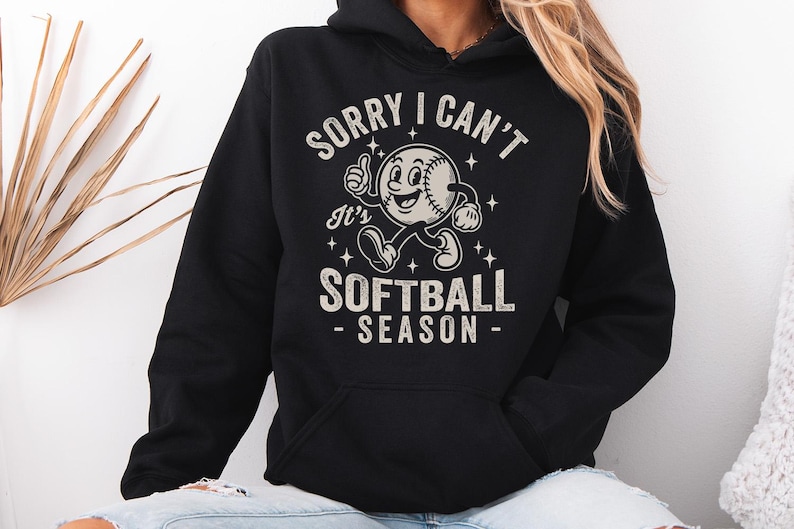 Op de afbeelding: Zwarte hoodie met een softbalafbeelding. De afbeelding toont een softbalpersonage met een duim omhoog en de tekst "SORRY I CAN'T It's SOFTBALL SEASON". De tekst en afbeelding zijn in wit en lichtgrijs.