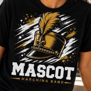 May include: Black t-shirt featuring a gold and black marching band hat with a feather plume. The word "MASCOT" is printed in bold white letters, with "MARCHING BAND" below. Gold and white accents complete the design.