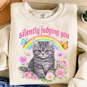 Pu&ograve; includere: Felpa color crema con un'illustrazione di un gatto tabby grigio scontroso e la scritta "silently judging you". Il design include un arcobaleno, farfalle e fiori, creando un'estetica stravagante e umoristica.