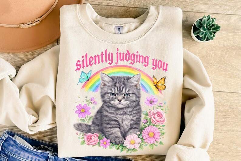 Pu&ograve; includere: Felpa color crema con un'illustrazione di un gatto tabby grigio scontroso e la scritta "silently judging you". Il design include un arcobaleno, farfalle e fiori, creando un'estetica stravagante e umoristica.