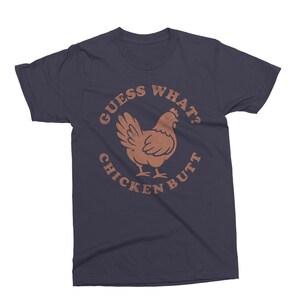 May include: Navy blue t-shirt with a brown graphic of a chicken and the text "GUESS WHAT? CHICKEN BUTT" in a circular design. The shirt is a crew neck style.