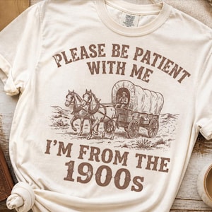 May include: Cream-colored t-shirt with a brown graphic of a covered wagon pulled by two horses. The text reads "PLEASE BE PATIENT WITH ME I'M FROM THE 1900s". A hat and a leather-bound journal are visible in the background.