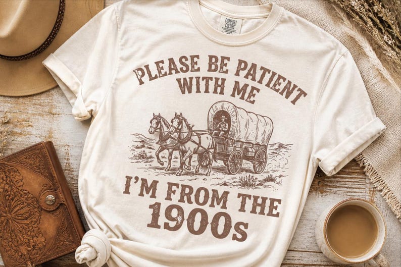 May include: Cream-colored t-shirt with a brown graphic of a covered wagon pulled by two horses. The text reads "PLEASE BE PATIENT WITH ME I'M FROM THE 1900s". A hat and a leather-bound journal are visible in the background.