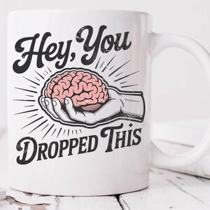 May include: White ceramic mug with a black and pink graphic. The graphic features a hand holding a pink brain with the text "Hey, You Dropped This" in a vintage style font. The mug has a curved handle.