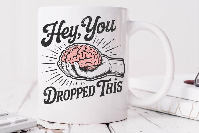 May include: White ceramic mug with a black and pink graphic. The graphic features a hand holding a pink brain with the text "Hey, You Dropped This" in a vintage style font. The mug has a curved handle.
