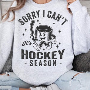 May include: Pale grey sweatshirt with the text "SORRY I CAN'T It's HOCKEY SEASON" in black. The graphic features a cartoon hockey puck with a hockey stick. The sweatshirt is a crew neck style.