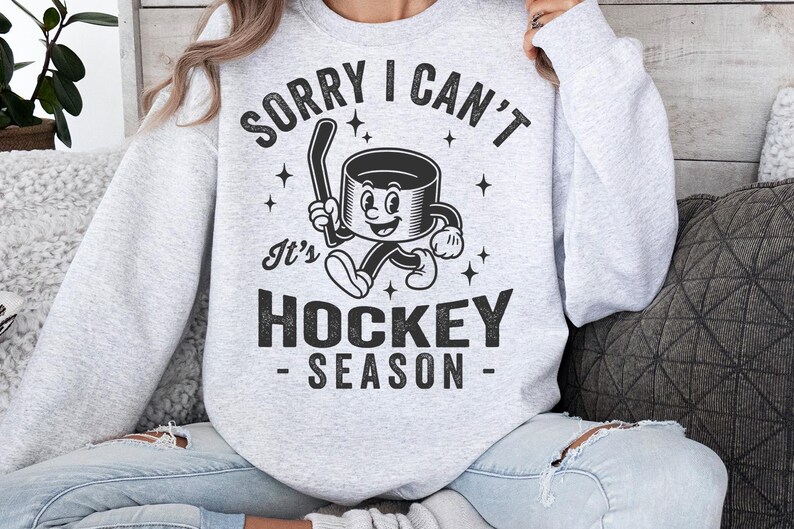 May include: Pale grey sweatshirt with the text "SORRY I CAN'T It's HOCKEY SEASON" in black. The graphic features a cartoon hockey puck with a hockey stick. The sweatshirt is a crew neck style.