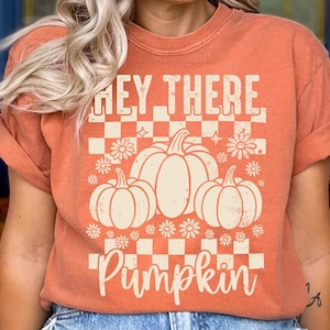 May include: Orange t-shirt featuring the text "Hey There Pumpkin" in a white, retro font. The design includes three pumpkins and floral accents, set against a checkered background. The shirt has short sleeves.