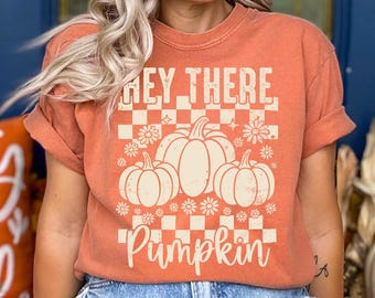 May include: Orange t-shirt with the text "Hey There Pumpkin" in a white, retro font. The design includes three pumpkins and floral accents, set against a checkered background. The shirt has short sleeves.