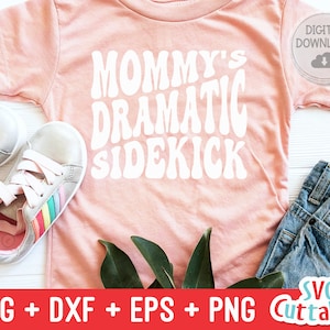 May include: Pink t-shirt with the text "Mommy's Dramatic Sidekick" in white, retro-style font. The shirt is laying on a white surface with a pair of rainbow shoelace sneakers and a pair of blue jeans.