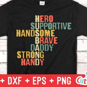 May include: A black t-shirt with a vintage-style design featuring the words "Hero", "Supportive", "Handsome", "Brave", "Daddy", "Strong", and "Handy" arranged in a crossword puzzle pattern.