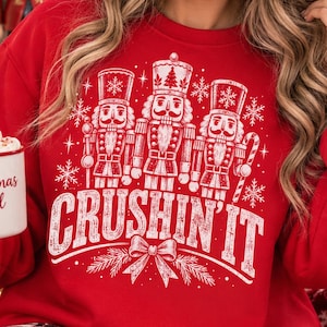 May include: A red crewneck sweatshirt featuring a white graphic print of three nutcracker figures, snowflakes, and a bow. The text "CRUSHIN' IT" is printed in large, distressed white capital letters across the center of the chest area.
