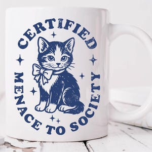 May include: White ceramic mug with a blue illustration of a cat wearing a bow tie, surrounded by the text "CERTIFIED MENACE TO SOCIETY" in a circular design. The mug has a curved handle and is on a white wooden surface.