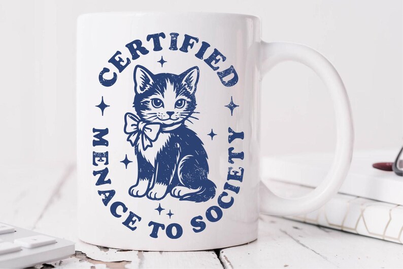 May include: White ceramic mug with a blue illustration of a cat wearing a bow tie, surrounded by the text "CERTIFIED MENACE TO SOCIETY" in a circular design. The mug has a curved handle and is on a white wooden surface.