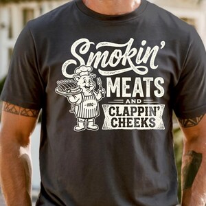 May include: Dark gray t-shirt with the text "Smokin' Meats and Clappin' Cheeks" in white. A cartoon pig chef holding a tray of ribs is featured in the design. The shirt is short-sleeved.