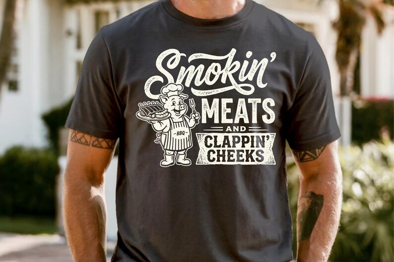 May include: Dark gray t-shirt with the text "Smokin' Meats and Clappin' Cheeks" in white. A cartoon pig chef holding a tray of ribs is featured in the design. The shirt is short-sleeved.