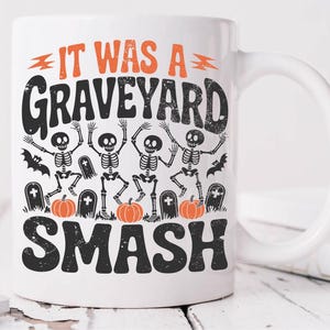 May include: White ceramic mug with a Halloween-themed design. The mug features the text "IT WAS A GRAVEYARD SMASH" in orange and black, along with illustrations of dancing skeletons, pumpkins, and tombstones. A perfect gift for Halloween.