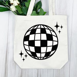 Puede incluir: Un bolso tote color crema con un dise&ntilde;o de bola de discoteca en blanco y negro y acentos de estrellas. El bolso tiene dos asas y est&aacute; hecho de una tela resistente. Flores blancas y hojas verdes son visibles en la parte superior.