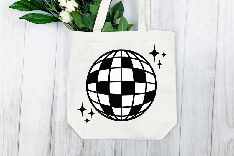 Puede incluir: Un bolso tote color crema con un dise&ntilde;o de bola de discoteca en blanco y negro y acentos de estrellas. El bolso tiene dos asas y est&aacute; hecho de una tela resistente. Flores blancas y hojas verdes son visibles en la parte superior.