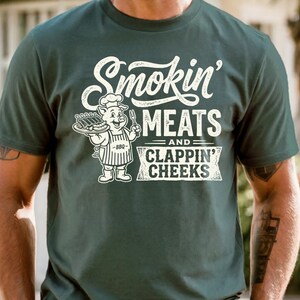 May include: Dark green t-shirt with the text "Smokin' MEATS and CLAPPIN' CHEEKS" in white. A cartoon pig chef holding a tray of ribs and a fork is also featured. The shirt is a casual wear item.