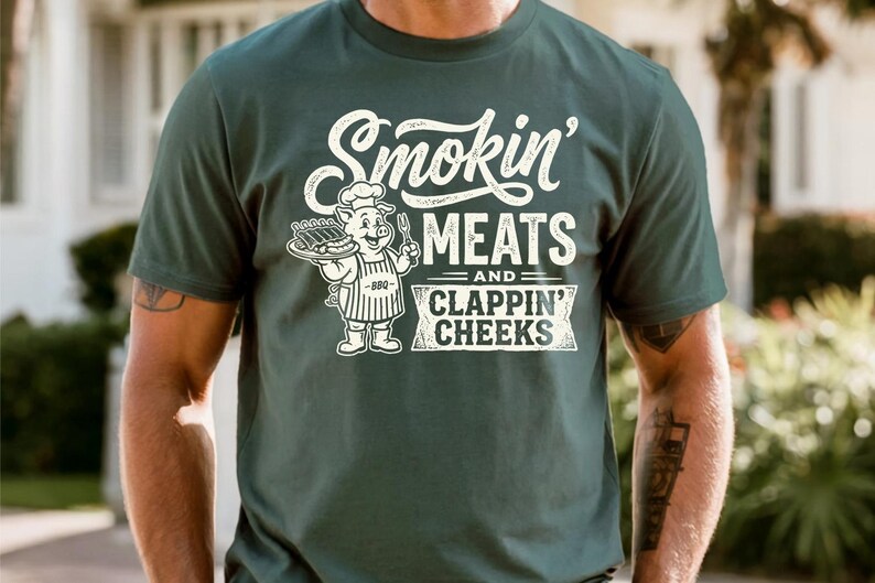 May include: Dark green t-shirt with the text "Smokin' MEATS and CLAPPIN' CHEEKS" in white. A cartoon pig chef holding a tray of ribs and a fork is also featured. The shirt is a casual wear item.