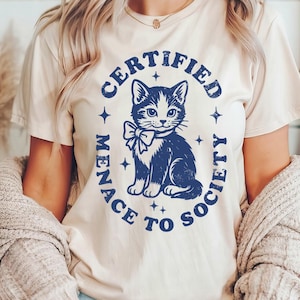 May include: Cream-colored t-shirt featuring a navy blue graphic of a cat with a bow tie, surrounded by the words "CERTIFIED MENACE TO SOCIETY". The design includes stars. The shirt is a casual wear item.