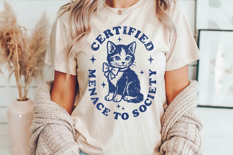 May include: Cream-colored t-shirt featuring a navy blue graphic of a cat with a bow tie, surrounded by the words "CERTIFIED MENACE TO SOCIETY". The design includes stars. The shirt is a casual wear item.