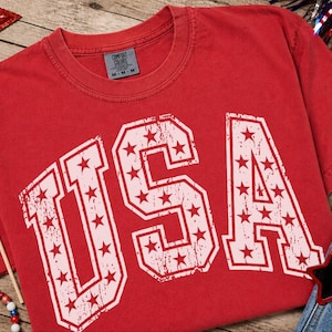 May include: Red t-shirt with the word "USA" in white, star-filled letters. The shirt is surrounded by patriotic decorations, including a small American flag, star-shaped sunglasses, and a red, white, and blue star.